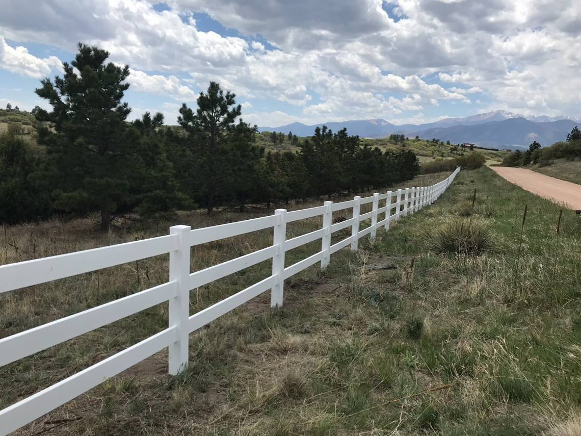 A long white fence stretches along a grassy field beside a dirt road, with trees and rolling hills in the background under a partly cloudy sky.
