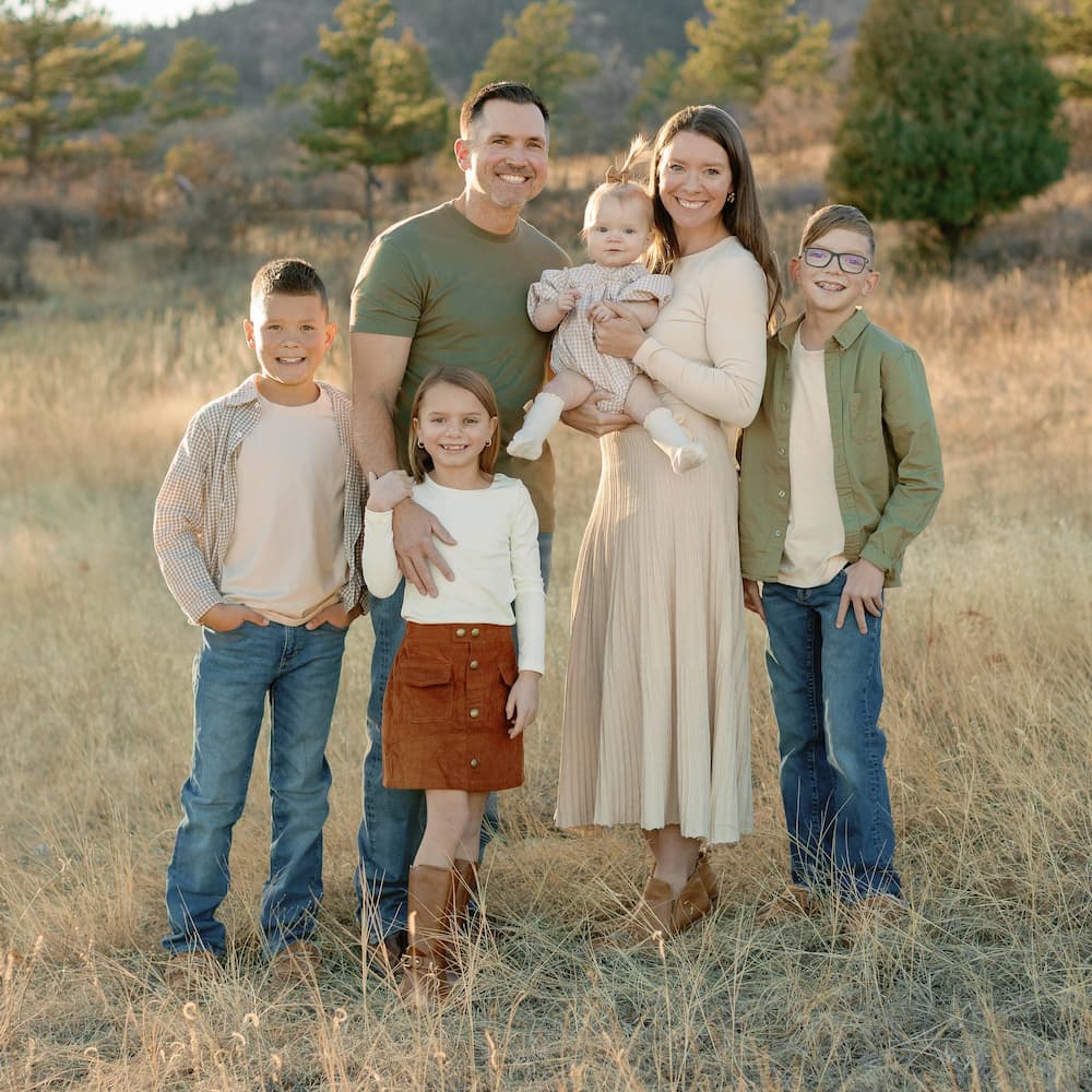 A family of six poses outdoors in a grassy field. Two adults stand in the center, the woman holding a baby. Three children stand around them, all smiling. Trees and hills are visible in the background under soft sunlight.