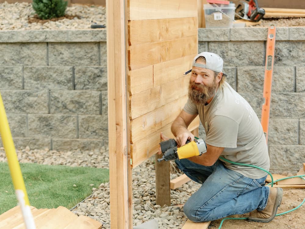A man wearing a gray shirt, jeans, and a backward cap kneels while building a wooden fence, holding a pneumatic nail gun. He is smiling at the camera. Tools and construction materials are visible in the background.