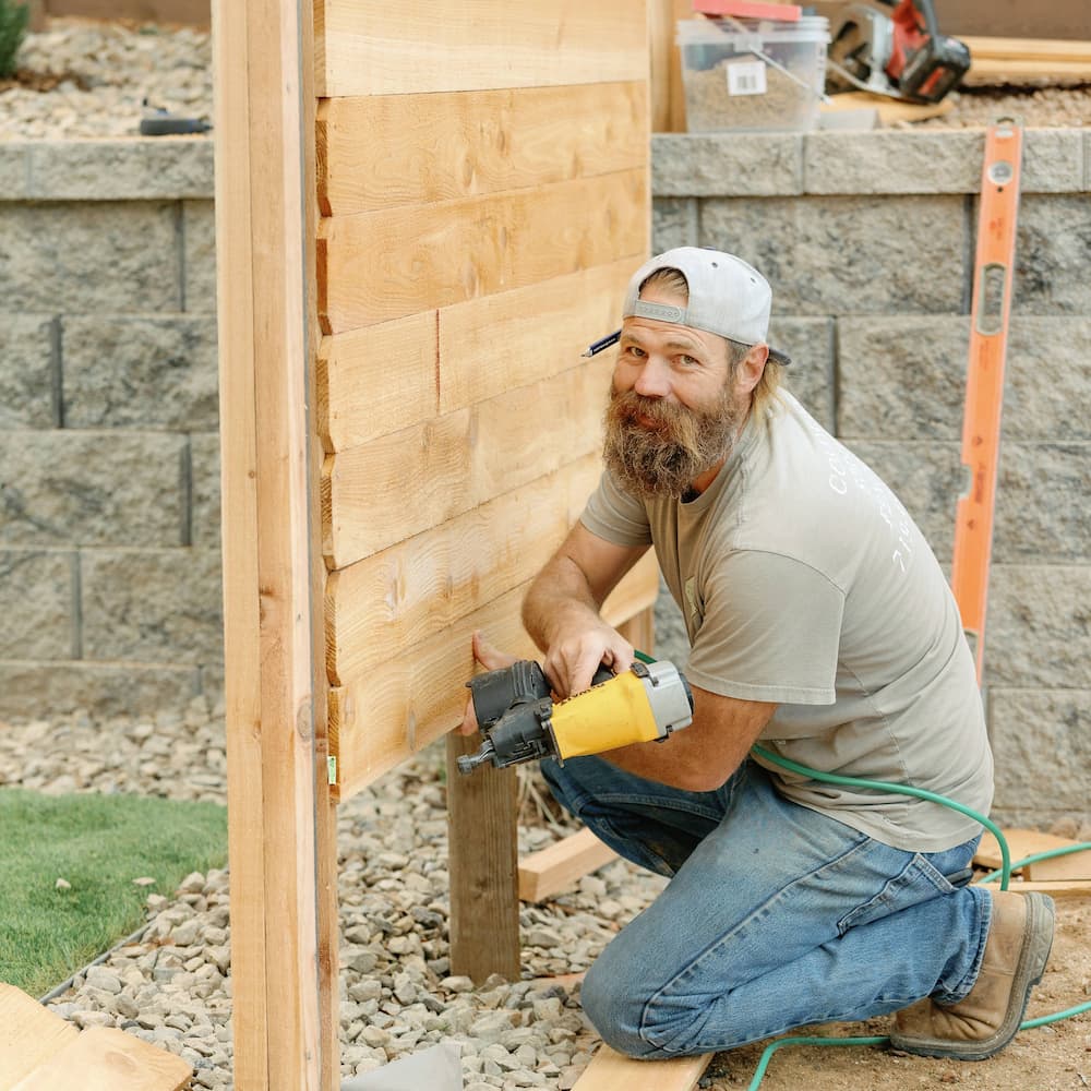 A man with a beard and a backwards cap kneels on the ground, using a nail gun to attach wooden boards to a fence frame. Construction tools and materials are scattered around him.
