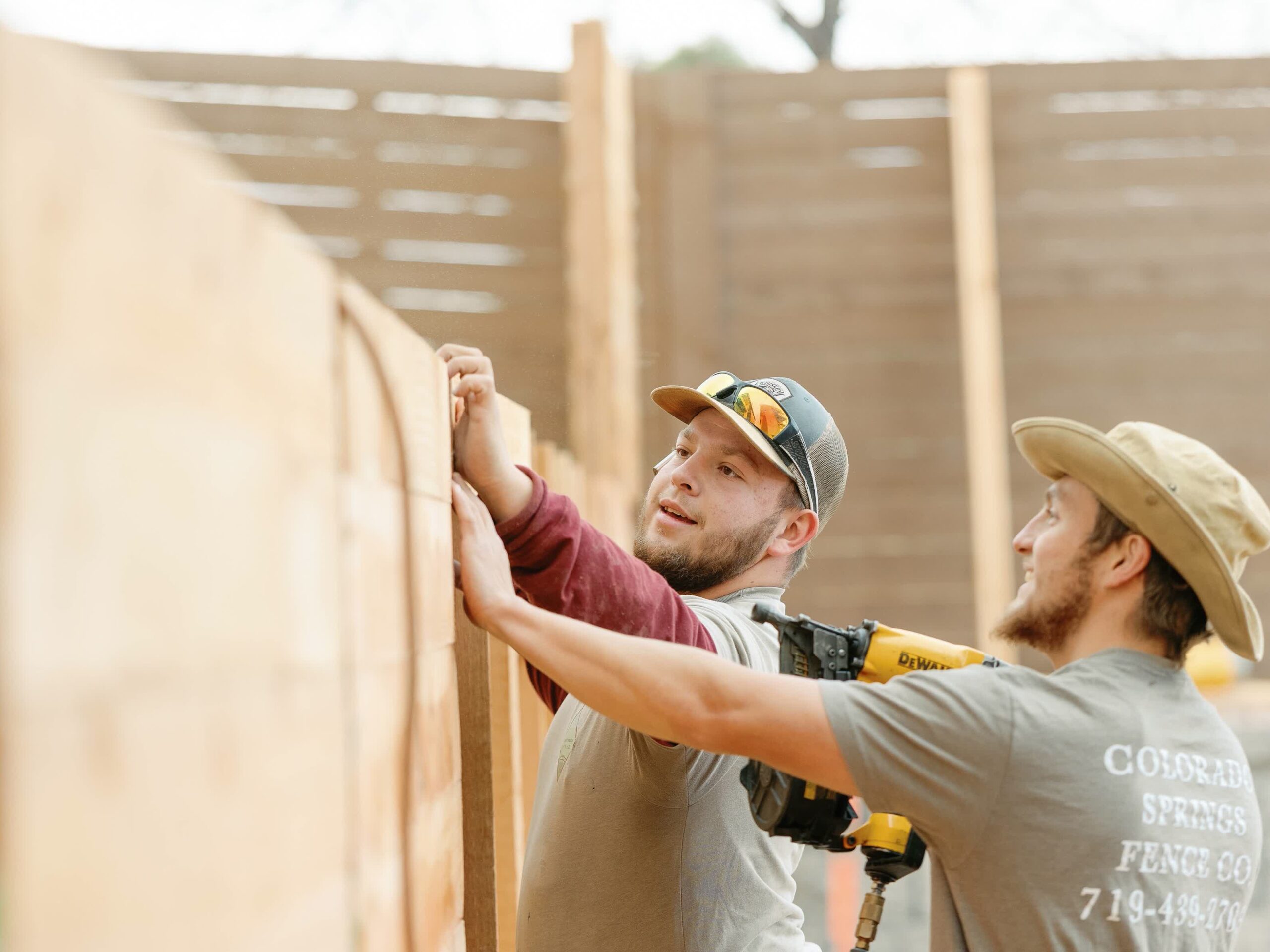 Two men wearing hats and gray shirts are installing wooden planks on a fence. One is holding the boards in place while the other uses a yellow nail gun. They appear focused on their construction task.