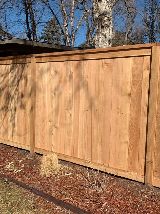 A new wooden privacy fence stands against a backdrop of leafless trees and a clear blue sky, with dry plants and mulch at the base of the fence.