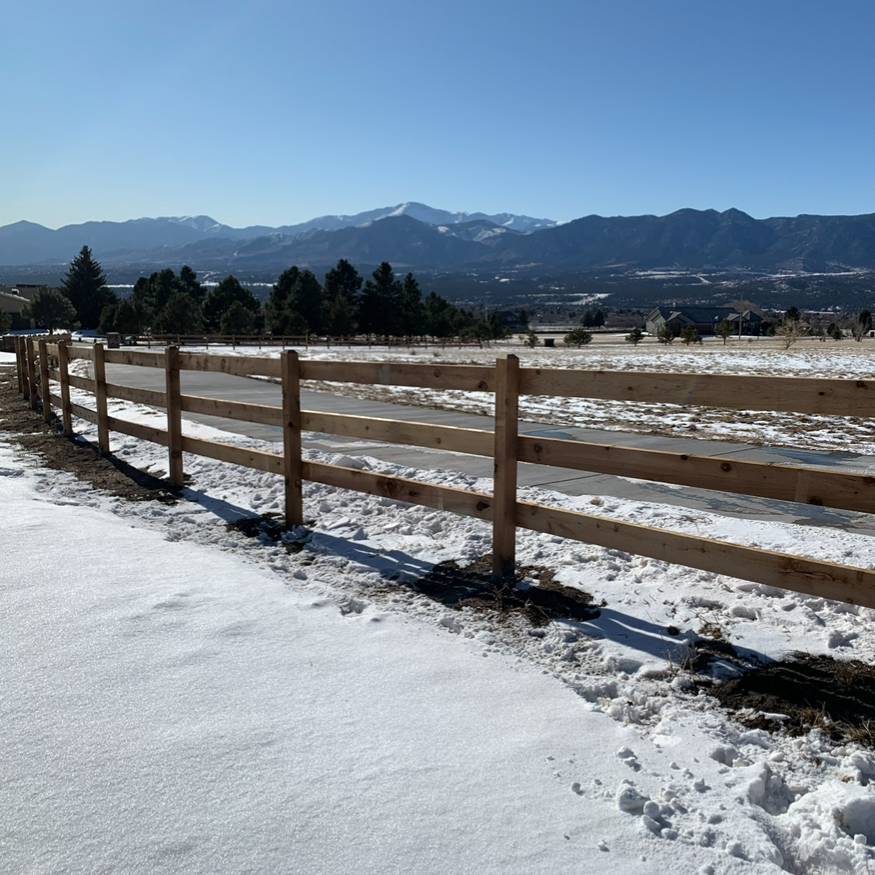 A wooden fence runs alongside a snow-covered field with scattered trees. In the background, tall mountains rise under a clear blue sky on a sunny winter day.