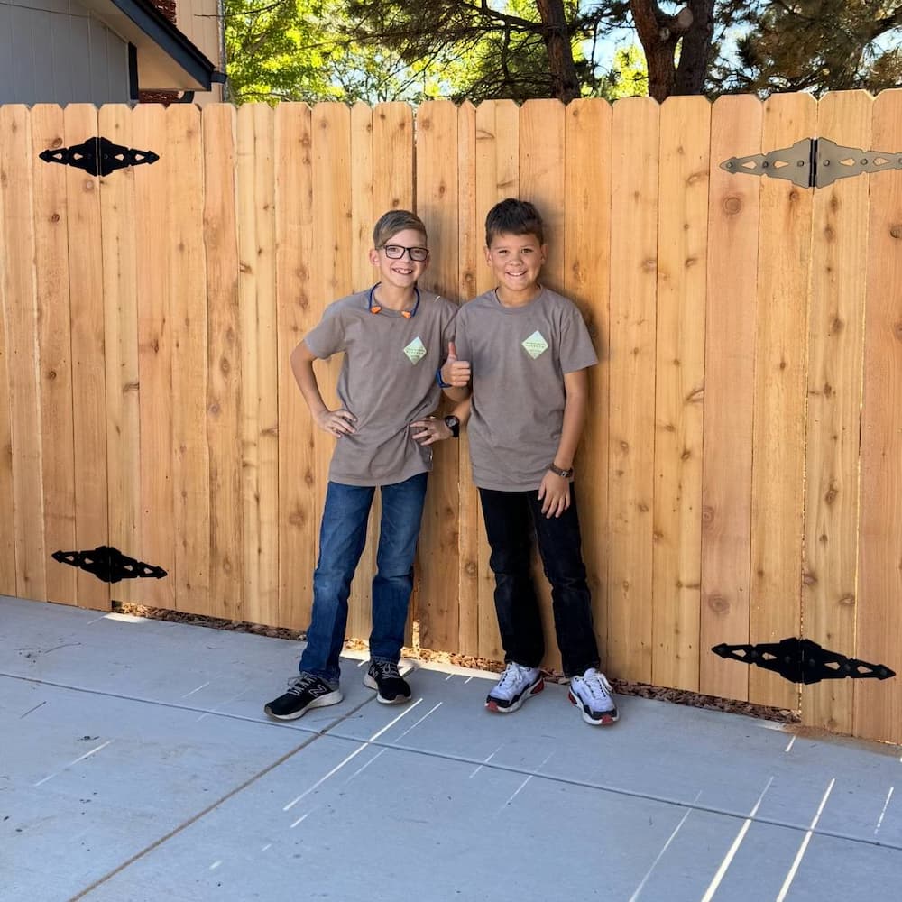 Two smiling kids wearing matching gray t-shirts and jeans stand in front of a tall wooden fence on a sunny day. Both have name tags, and the ground is paved with concrete.