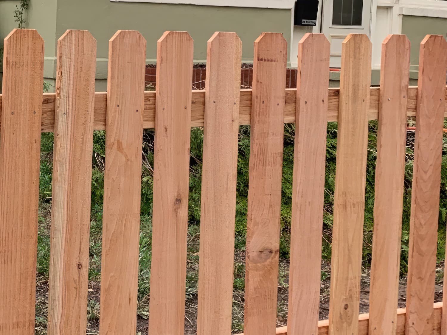 A close-up view of a new, light-colored wooden picket fence in front of a house with green siding and a white door. Grass and part of a brick step are visible behind the fence.