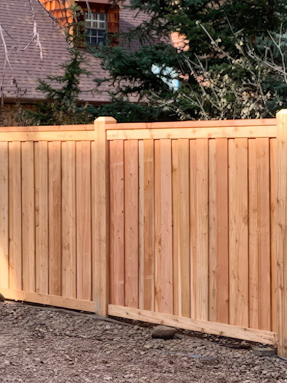 A new wooden fence with vertical panels stands on rocky ground, with part of a house and green trees visible in the background.
