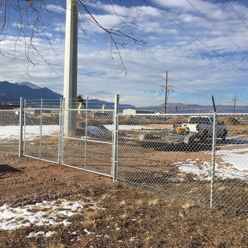 A chain-link fence with a gate encloses a utility pole and a pickup truck on a partly snowy, gravel-covered area. Mountains and a partly cloudy sky are visible in the background.