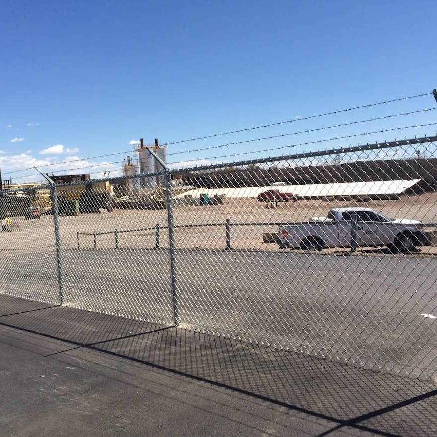 A barbed-wire chain-link fence separates an empty paved lot from an industrial area with factory buildings and a white pickup truck parked inside. The sky is clear and blue.