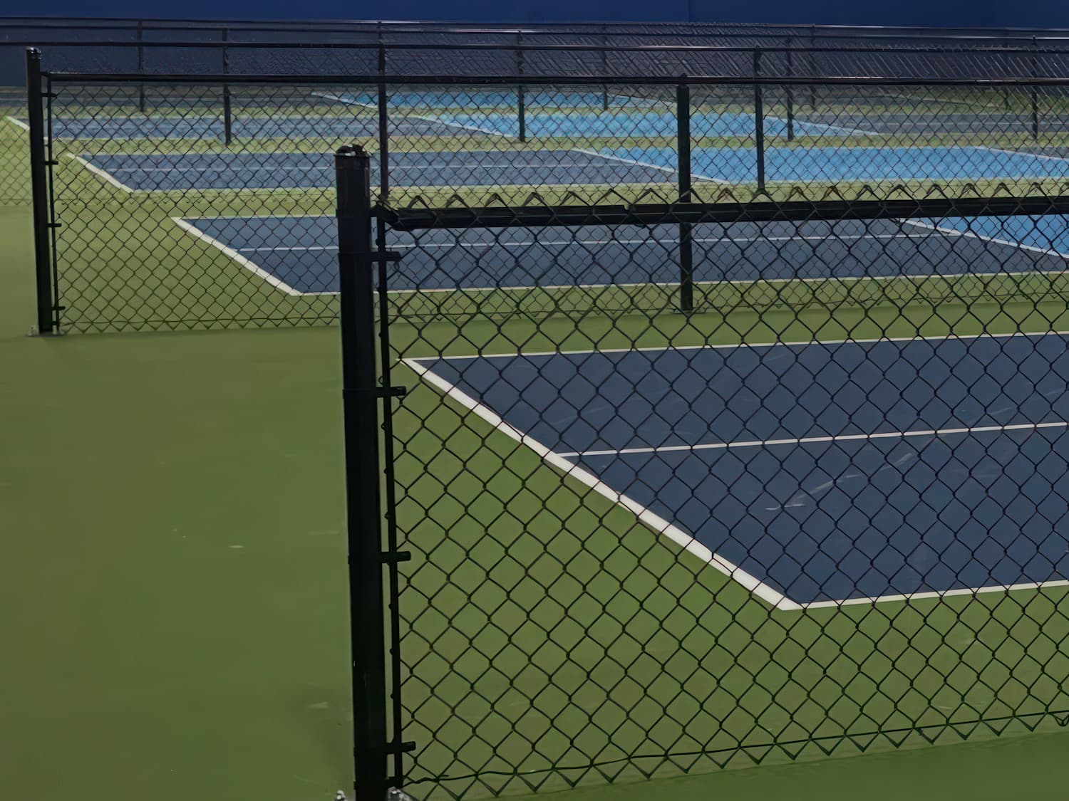 A chain-link fence separates several empty outdoor tennis courts with blue and green surfaces under clear daylight. The courts are lined up in rows, and no people are present.
