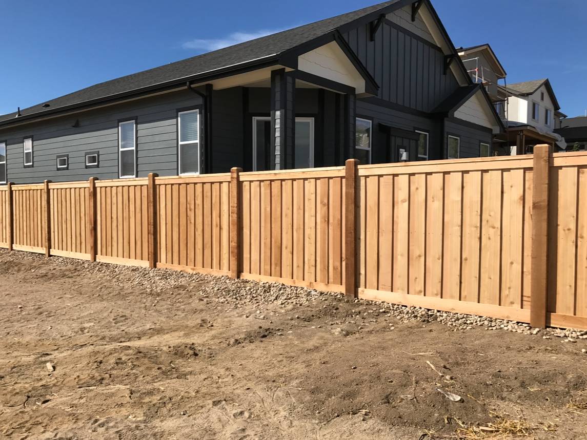 A modern house with dark gray siding stands behind a newly built wooden privacy fence on a dirt lot under a clear blue sky.