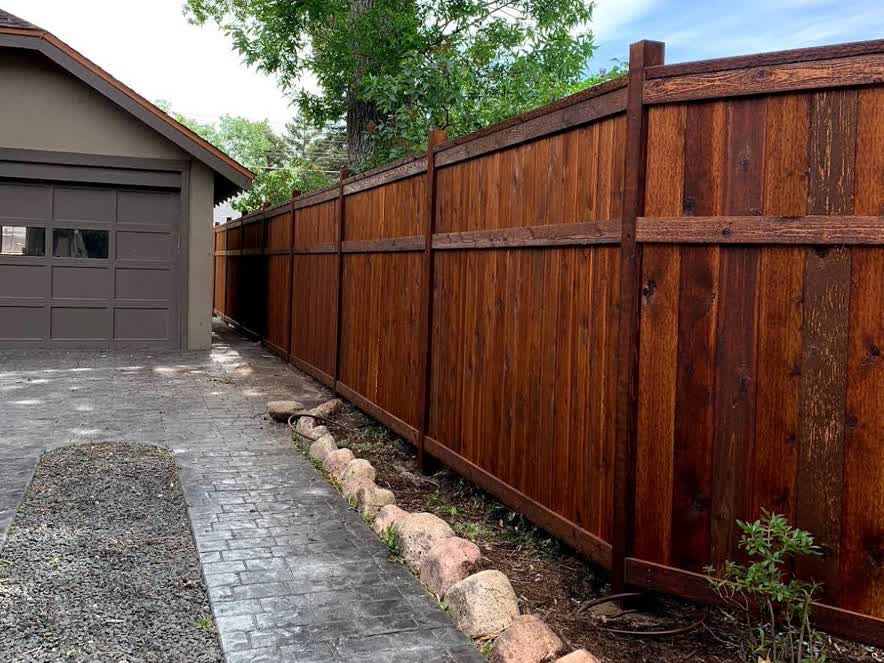 A tall, dark brown wooden privacy fence runs alongside a paved driveway and a small garden with rocks and sparse plants, next to a house with a gray garage door. Trees and a blue sky are visible in the background.
