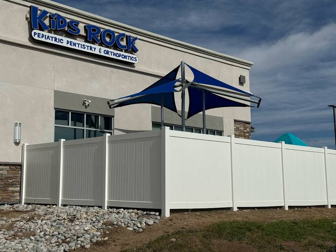 A white privacy fence surrounds an area outside a building with a sign that reads "Kids Rock Pediatric Dentistry & Orthodontics." A blue and white shade structure is visible behind the fence. The sky is partly cloudy.