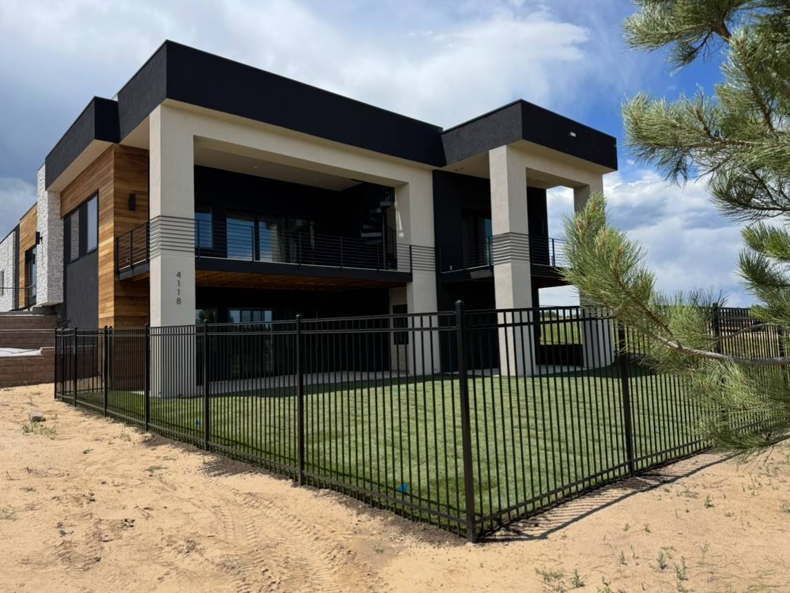 A modern two-story house with a flat roof, large windows, and a balcony. The exterior is black, white, and wood, with a black metal fence surrounding a green lawn. A tree and sandy ground are in the foreground.
