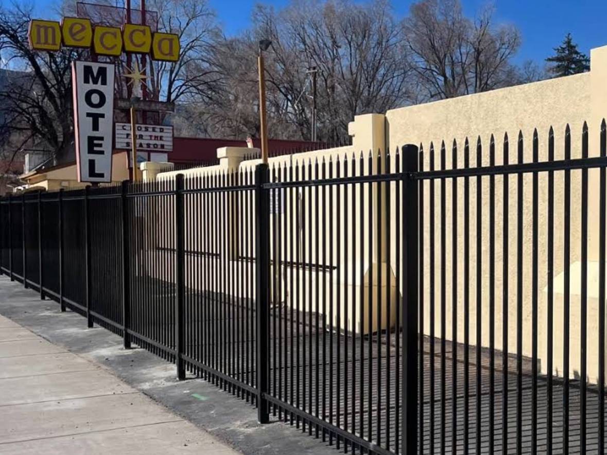 A black metal fence runs alongside a sidewalk in front of a beige building, with a vintage "Mecca Motel" sign and leafless trees visible in the background under a clear blue sky.