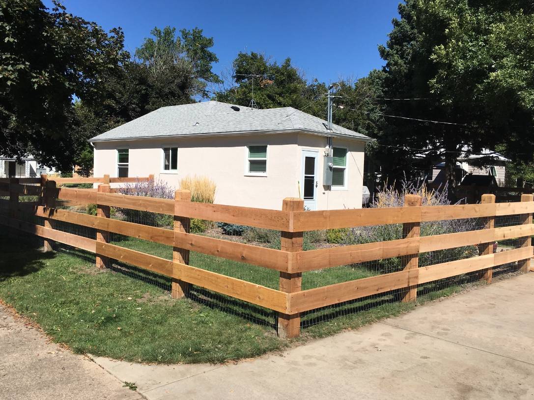 A small, white house with a gray roof sits behind a new wooden fence on a sunny day. The yard is landscaped with grass, bushes, and ornamental grasses, surrounded by trees and a clear blue sky.