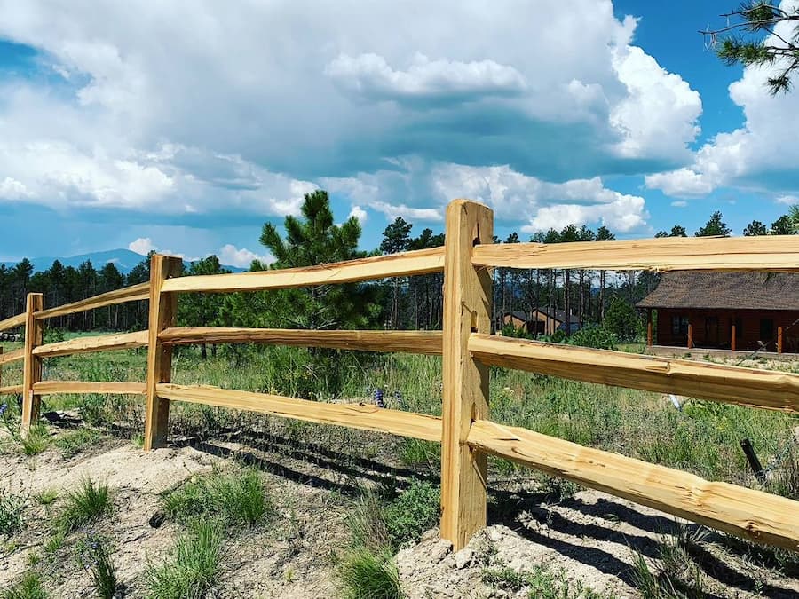 A wooden split rail fence runs diagonally across a grassy, slightly sandy field with scattered trees and a house in the background under a blue sky with white clouds.