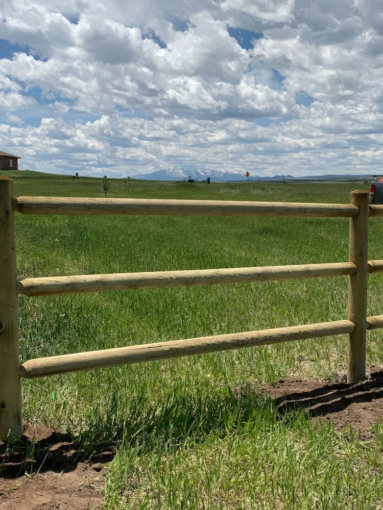 A wooden fence with three horizontal rails stands in front of a green grassy field under a partly cloudy sky. Mountains are visible in the distance.