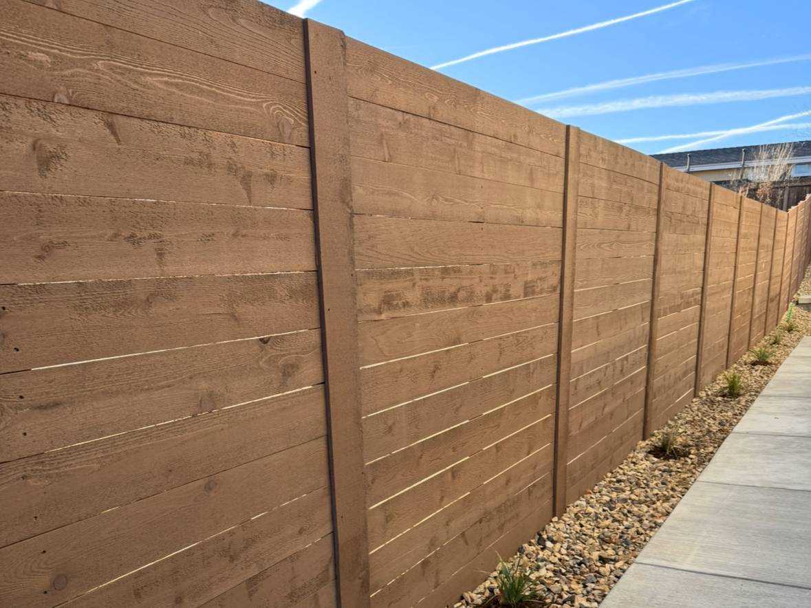 A long wooden privacy fence lines a sidewalk bordered by small rocks and plants, with clear blue sky and contrail streaks overhead.