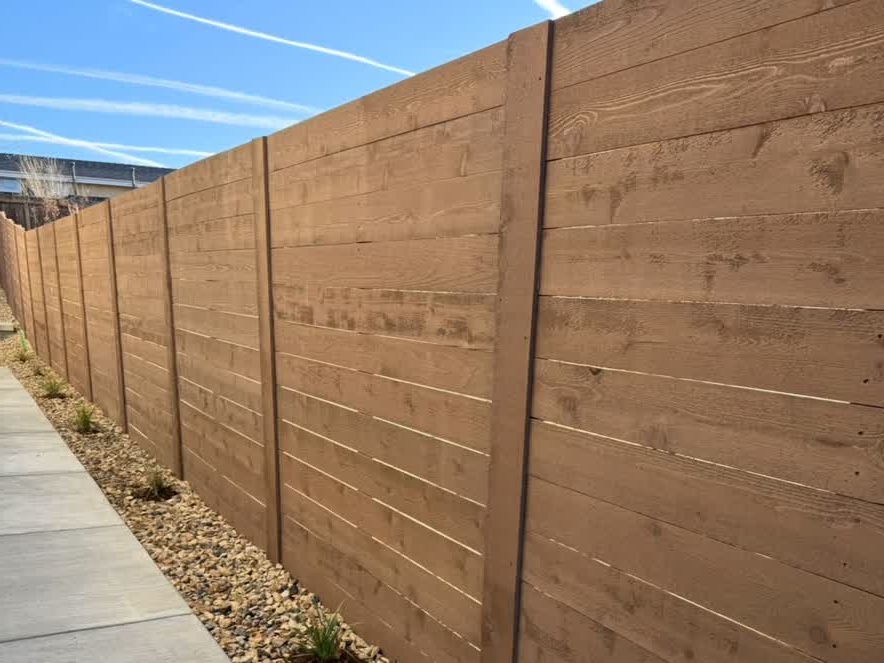A tall wooden fence runs alongside a sidewalk bordered with rocks and small plants. The sky above is blue with visible contrails.