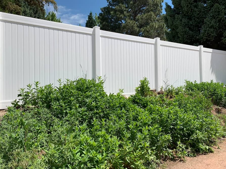 A tall white vinyl privacy fence runs along a yard with green leafy shrubs and plants growing in front of it under a partly cloudy sky.
